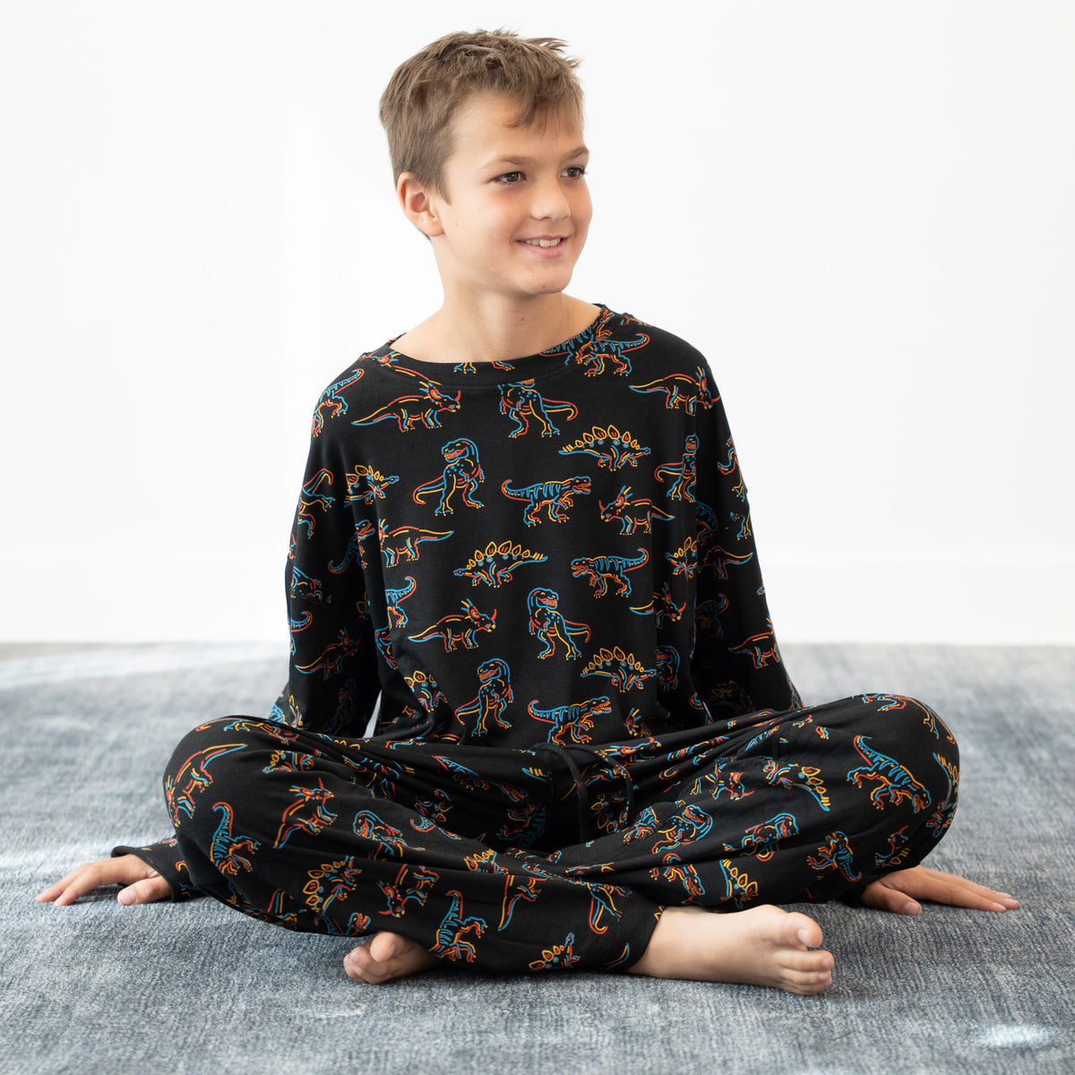 A young boy sitting cross-legged on a gray textured floor against a white wall, wearing a black long-sleeve pajama top and matching pants with a colorful dinosaur print.
