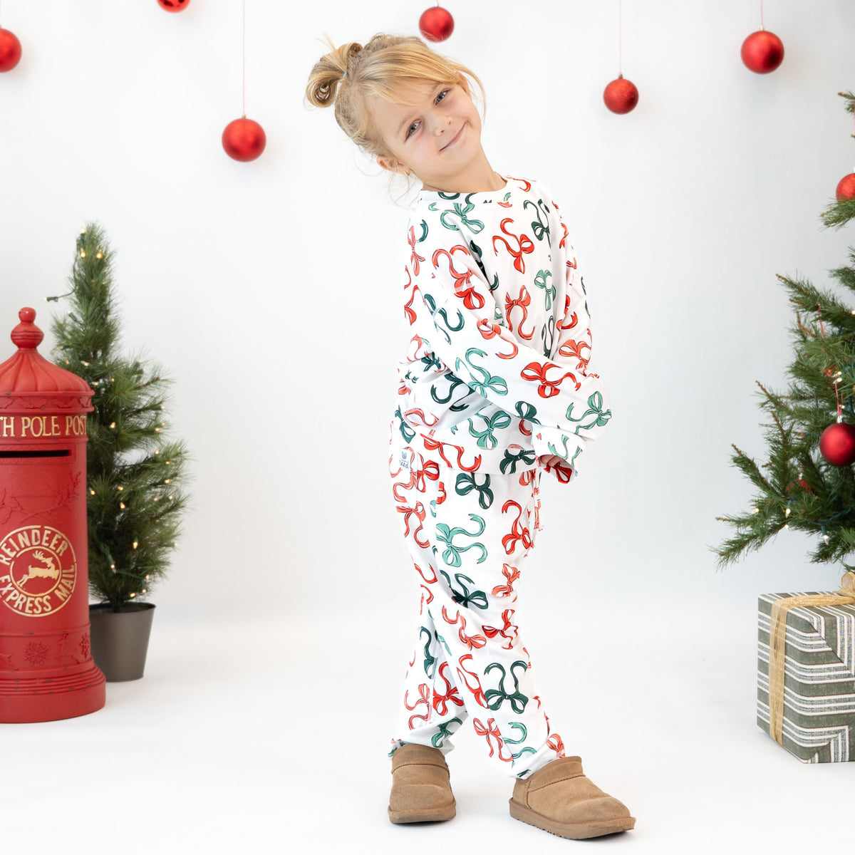 A young girl wearing a white long-sleeve pajama set with a red and green holiday pattern, standing in a festive studio decorated with Christmas trees, a red post box, and hanging ornaments.