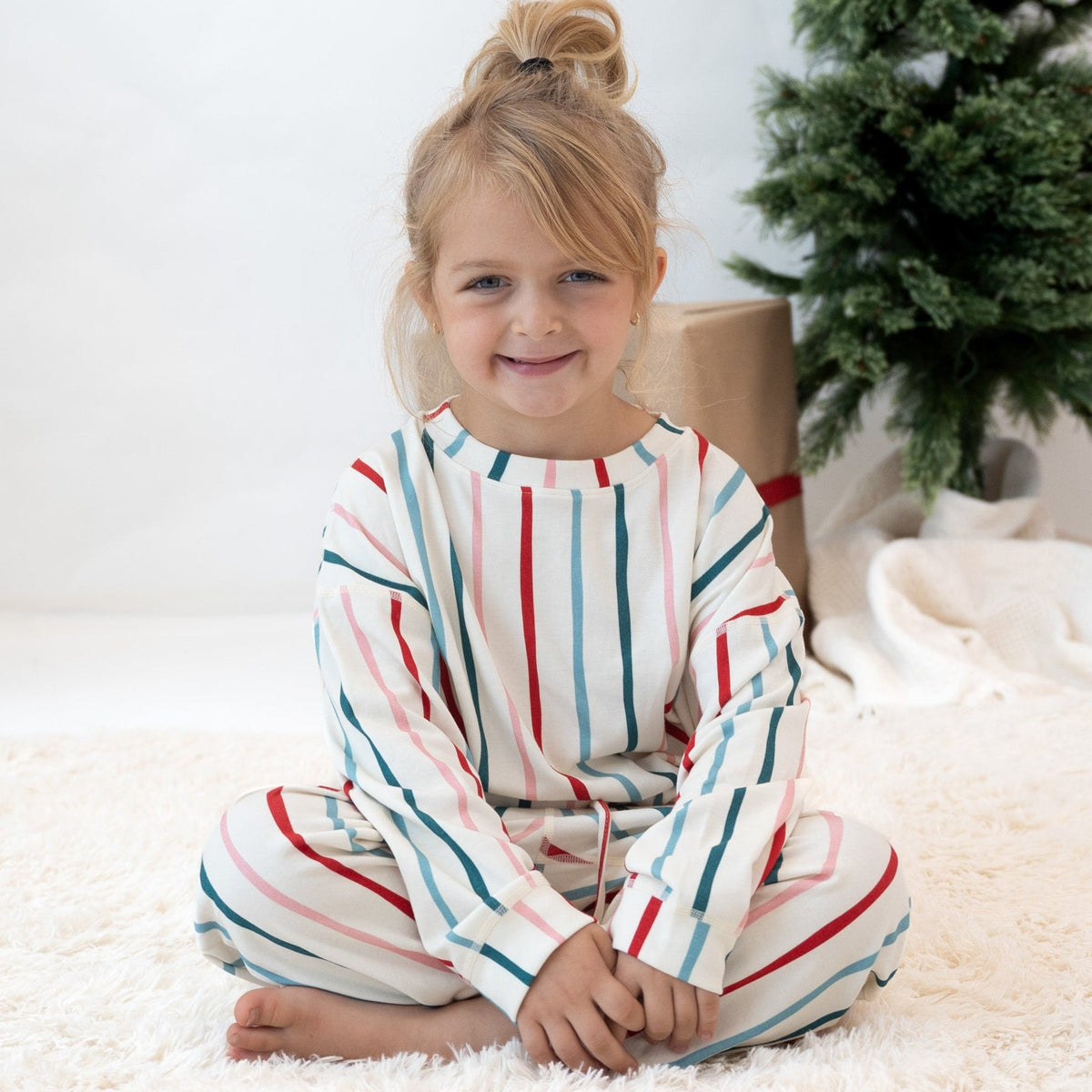 A young girl with blonde hair in a bun sits cross-legged on a white shag rug, wearing a matching long-sleeve pajama set with vertical stripes in red, teal, and light blue on an off-white base. A wrapped gift box and a Christmas tree are visible in the background.