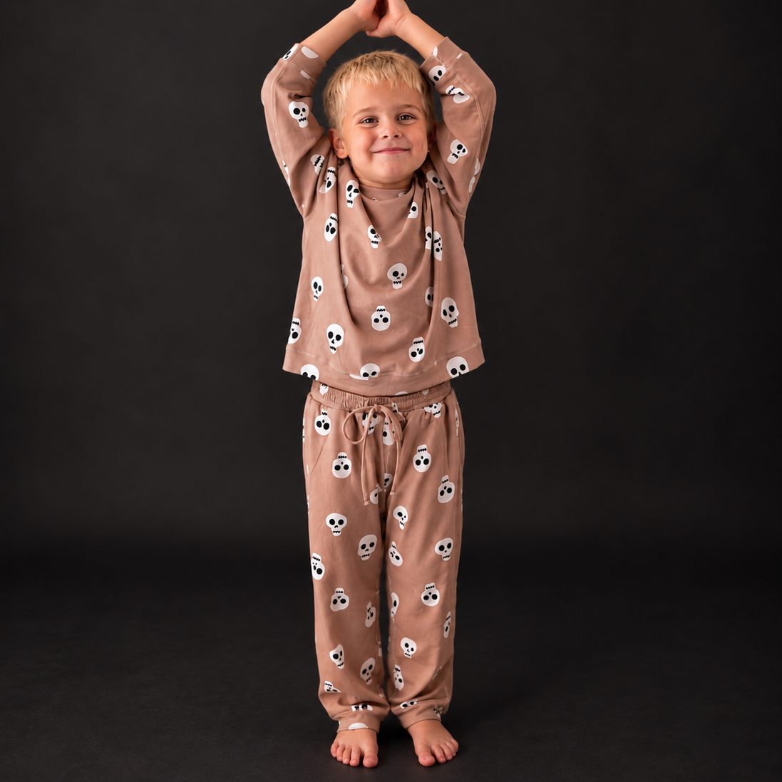 A young child stands barefoot against a dark studio background, wearing a matching long-sleeve pajama set in a warm taupe-brown color with a repeating white and black skull print.