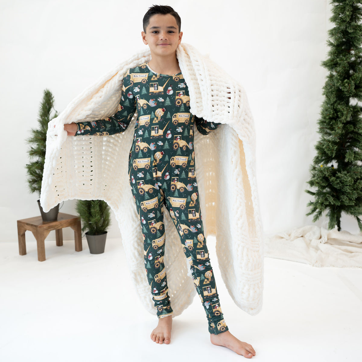 A child stands barefoot in a studio wearing a dark green long-sleeve pajama set with a tractor print, while holding a chunky white knit blanket. The background is white with small potted trees and a Christmas tree prop.