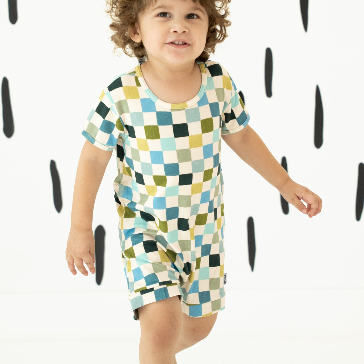 A toddler wearing a short-sleeve romper with a colorful geometric checkered pattern in blue, green, yellow, and white, set against a white background with black raindrop shapes.
