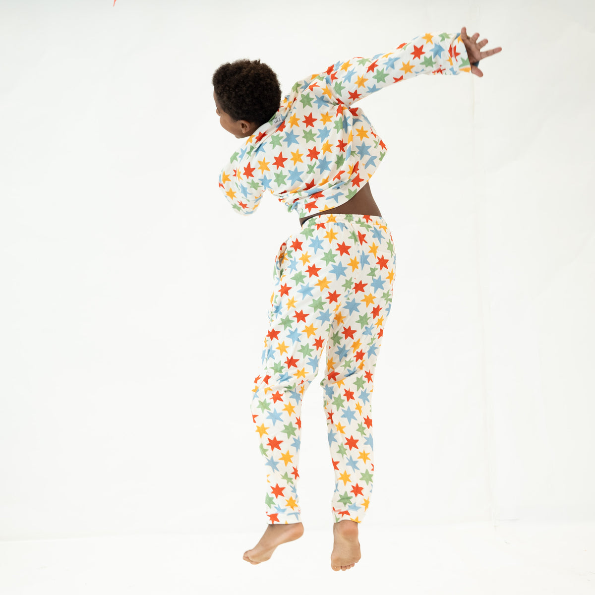 A person wearing a matching long-sleeve crop top and jogger pants set with a colorful star pattern, shown from the back while jumping against a plain white studio background.