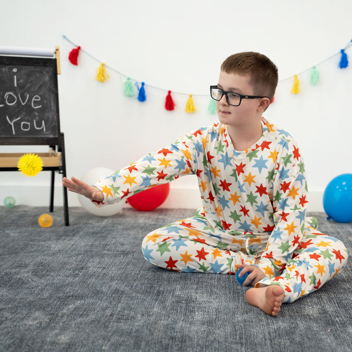 A young boy wearing glasses sits cross-legged on a gray rug, wearing a white long-sleeve pajama set covered in colorful star patterns. The room has a white wall decorated with colorful tassels, a small chalkboard easel, and balloons.