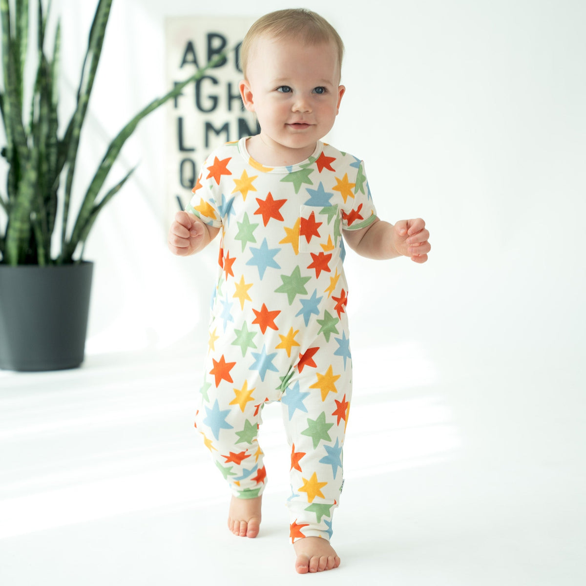 A baby walking barefoot in a white footless one-piece jumpsuit covered in colorful star patterns, standing in a bright room with a potted snake plant in the background.