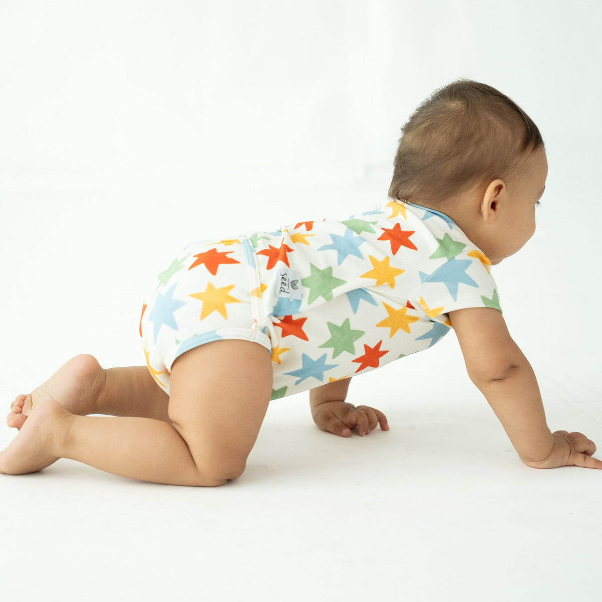 A baby crawling on a white studio floor wearing a white short-sleeve bodysuit covered in colorful star patterns.