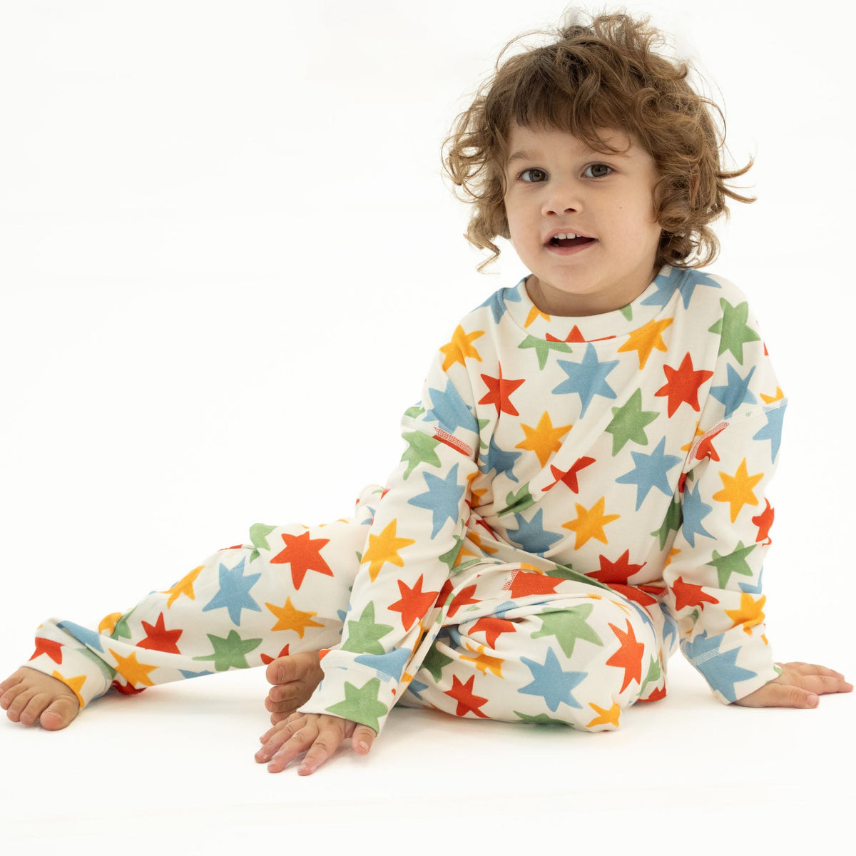 A toddler with curly brown hair sits cross-legged on a white studio background wearing a cream-colored long-sleeve pajama set covered in colorful star prints in blue, green, orange, and yellow.