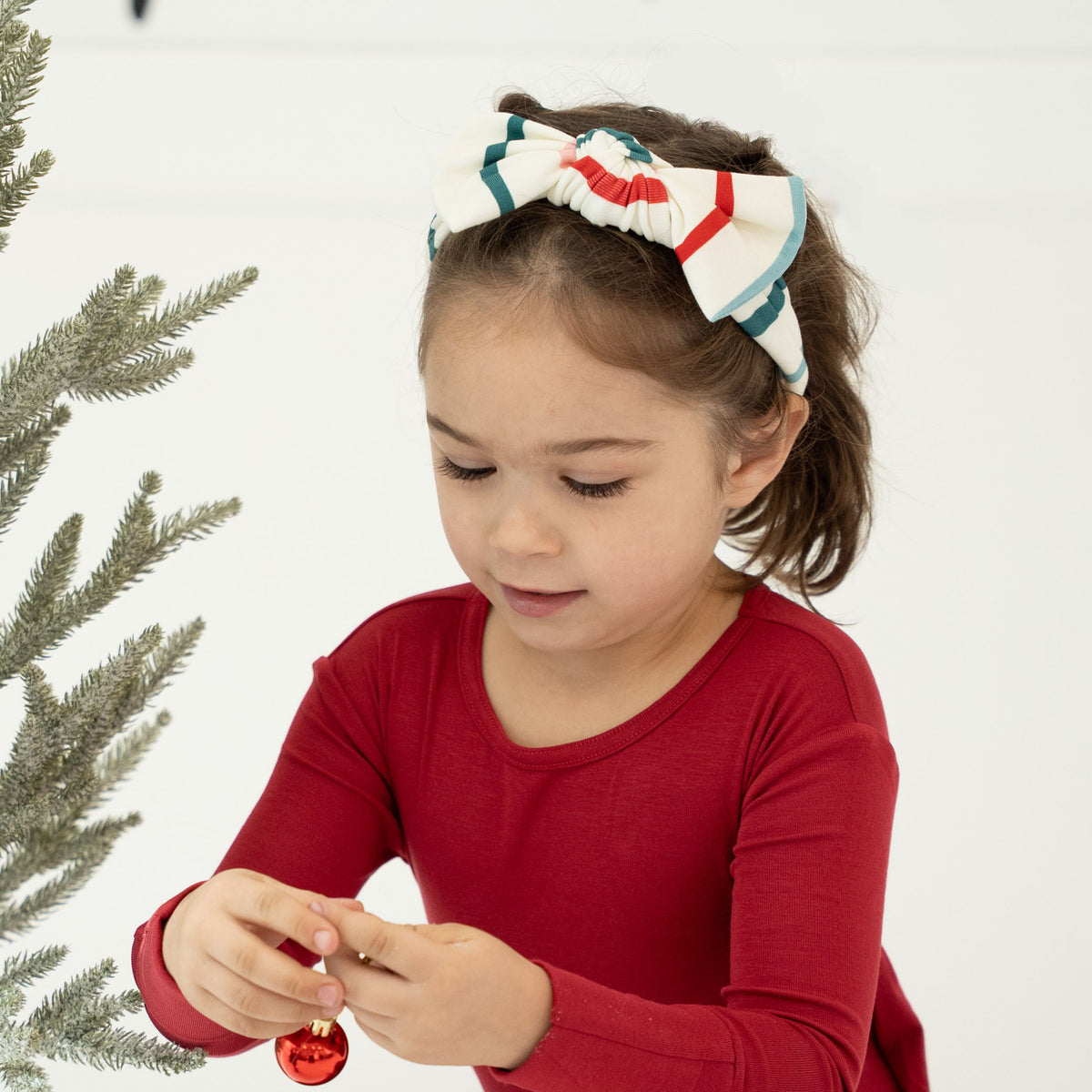 A young girl wearing a red long-sleeve shirt and a white bow headband with red and green stripes, looking down while holding a small red ornament. A Christmas tree branch is visible on the left against a white background.
