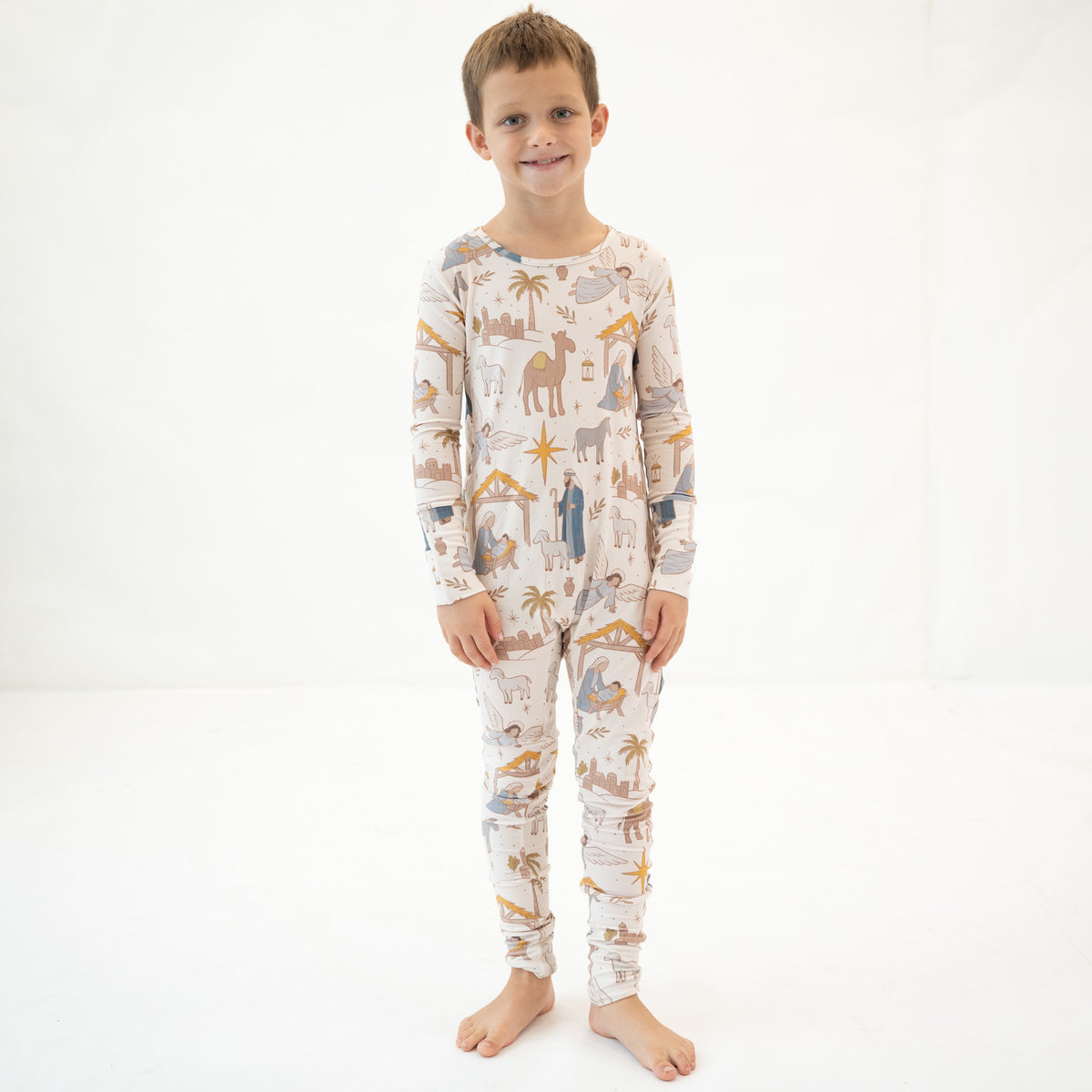 A young boy stands barefoot against a seamless white studio background, wearing a long-sleeve one-piece pajama set with an all-over desert-themed print featuring camels, palm trees, and small figures in muted blue, tan, and brown tones.