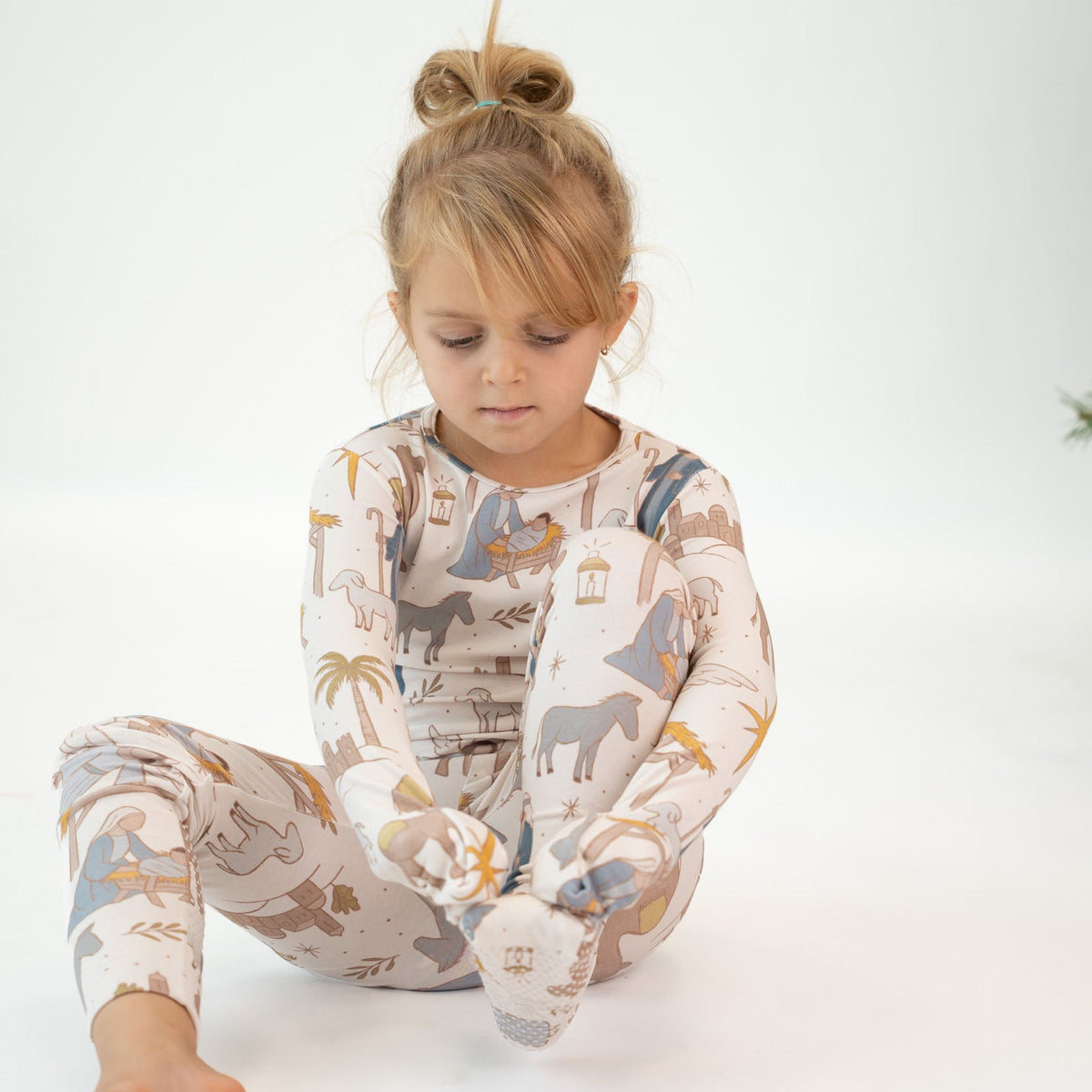 A young girl with blonde hair in a bun sits on a white studio floor wearing a matching long-sleeve pajama set with a safari animal print in beige, blue, and brown.