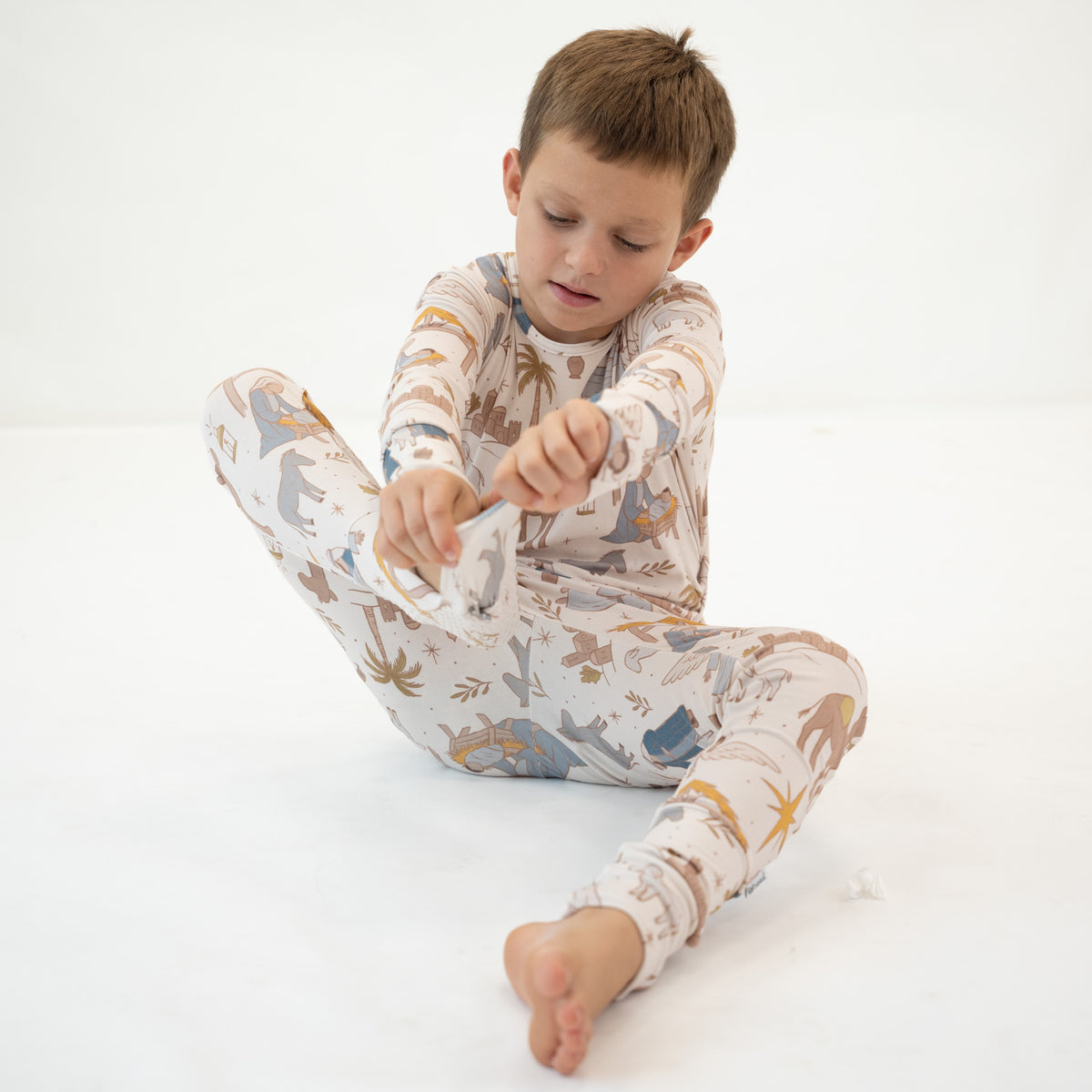 A young boy sitting cross-legged on a white studio floor, wearing a long-sleeve pajama set with a dinosaur and safari animal print in beige, blue, and brown tones.