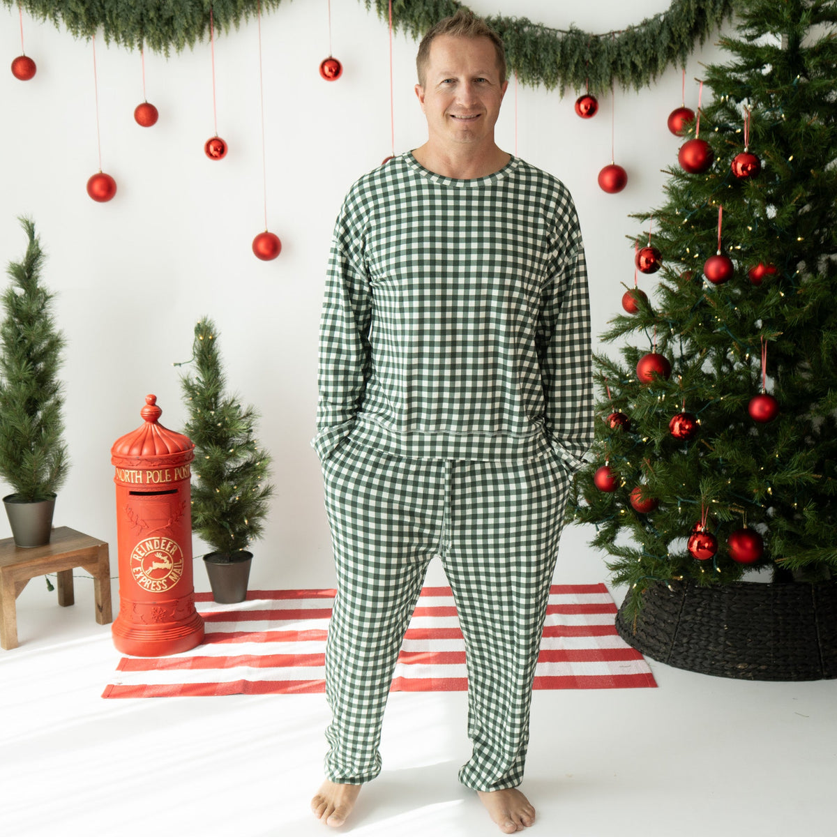 A man wearing a green and white gingham check long-sleeve pajama set stands barefoot in a decorated Christmas room with a tree, garland, and red ornaments.