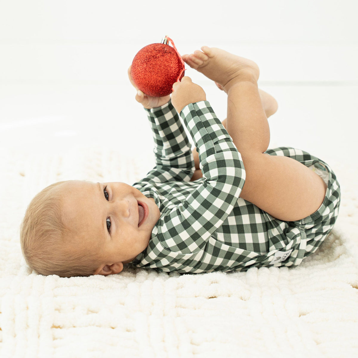 A baby lying on a white textured blanket, wearing a green and white gingham long-sleeve romper and playfully reaching for a red glittery ornament.