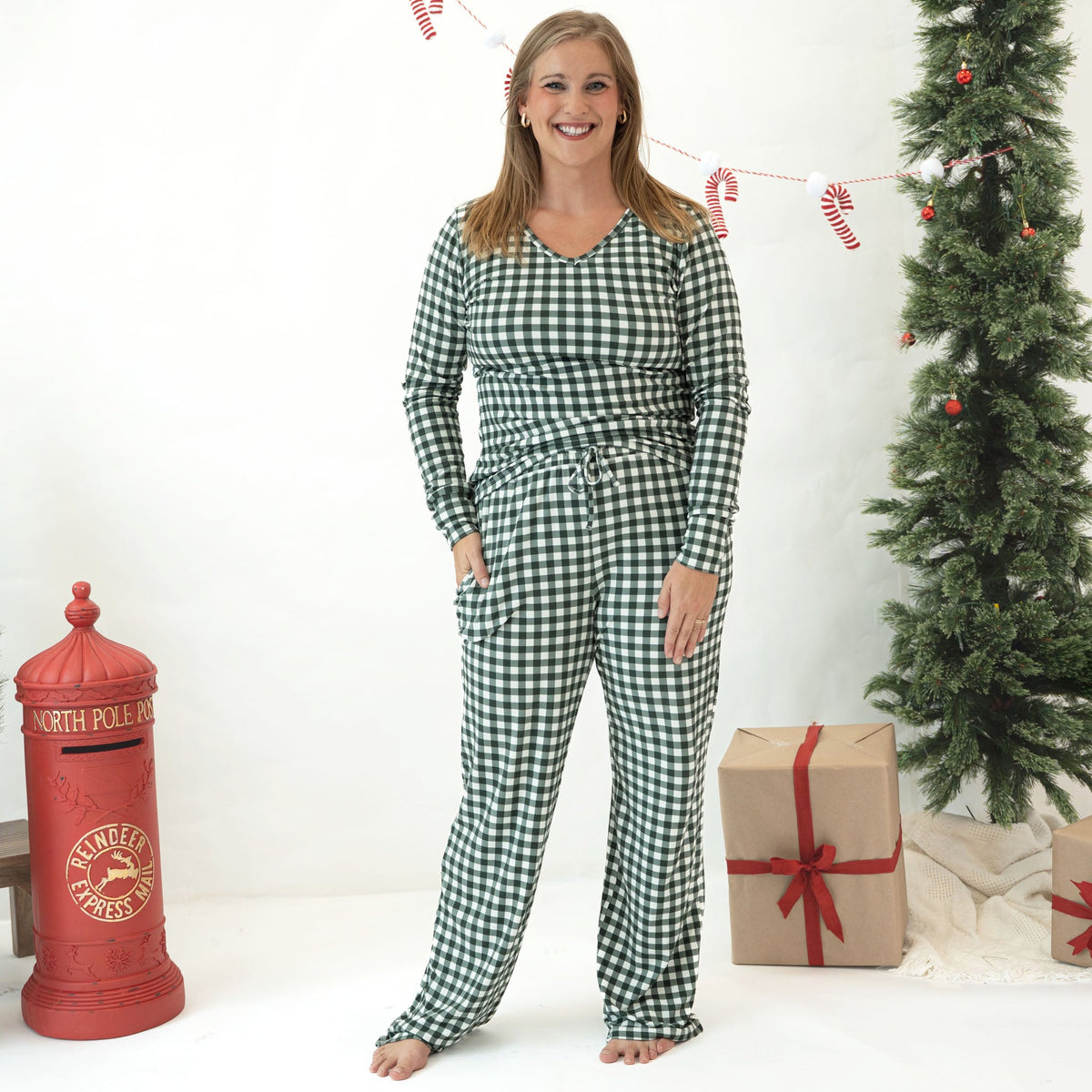 A woman stands barefoot in a holiday-themed room wearing a green and white gingham long-sleeve pajama set. The scene includes a red vintage-style North Pole mailbox, a decorated Christmas tree, wrapped gifts, and candy cane garland.