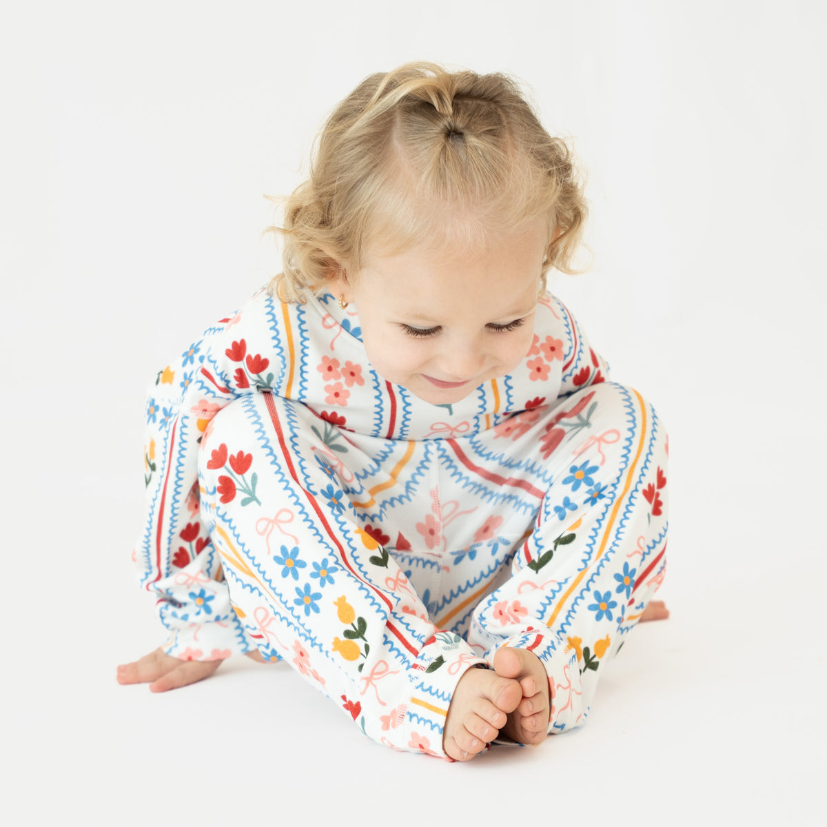 A toddler girl with blonde hair in a small ponytail sits cross-legged on a white studio background, wearing a long-sleeve pajama top and matching pants with a colorful floral and geometric pattern.