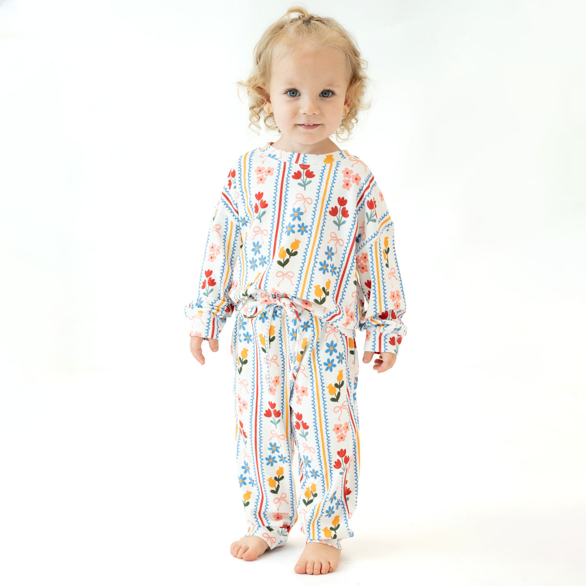A toddler with blonde curly hair stands barefoot against a white studio background, wearing a long-sleeve matching pajama set with a colorful floral and striped pattern.