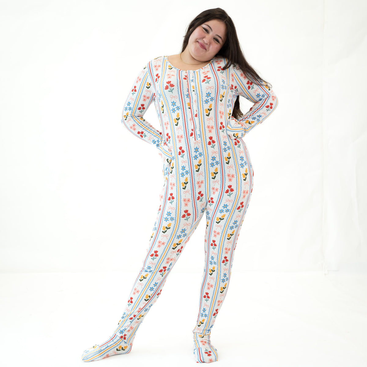 A woman modeling a one-piece footed pajama suit with a colorful vertical stripe pattern featuring cartoon birds and flowers on a white background.