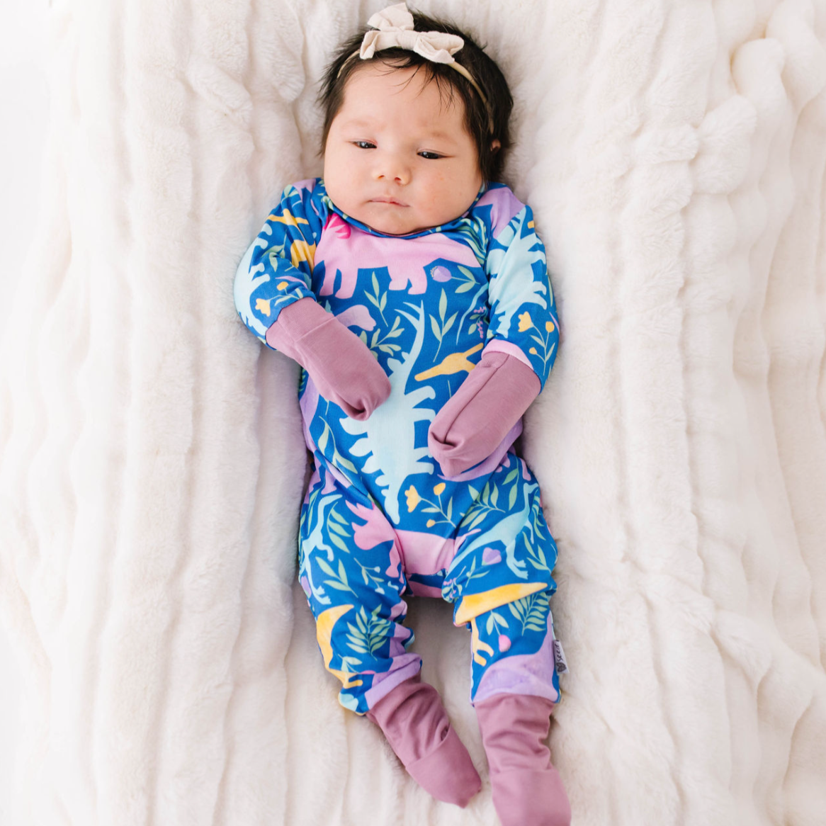 A baby lying on a soft surface wearing a long-sleeved romper with a dinosaur pattern in shades of blue and purple.