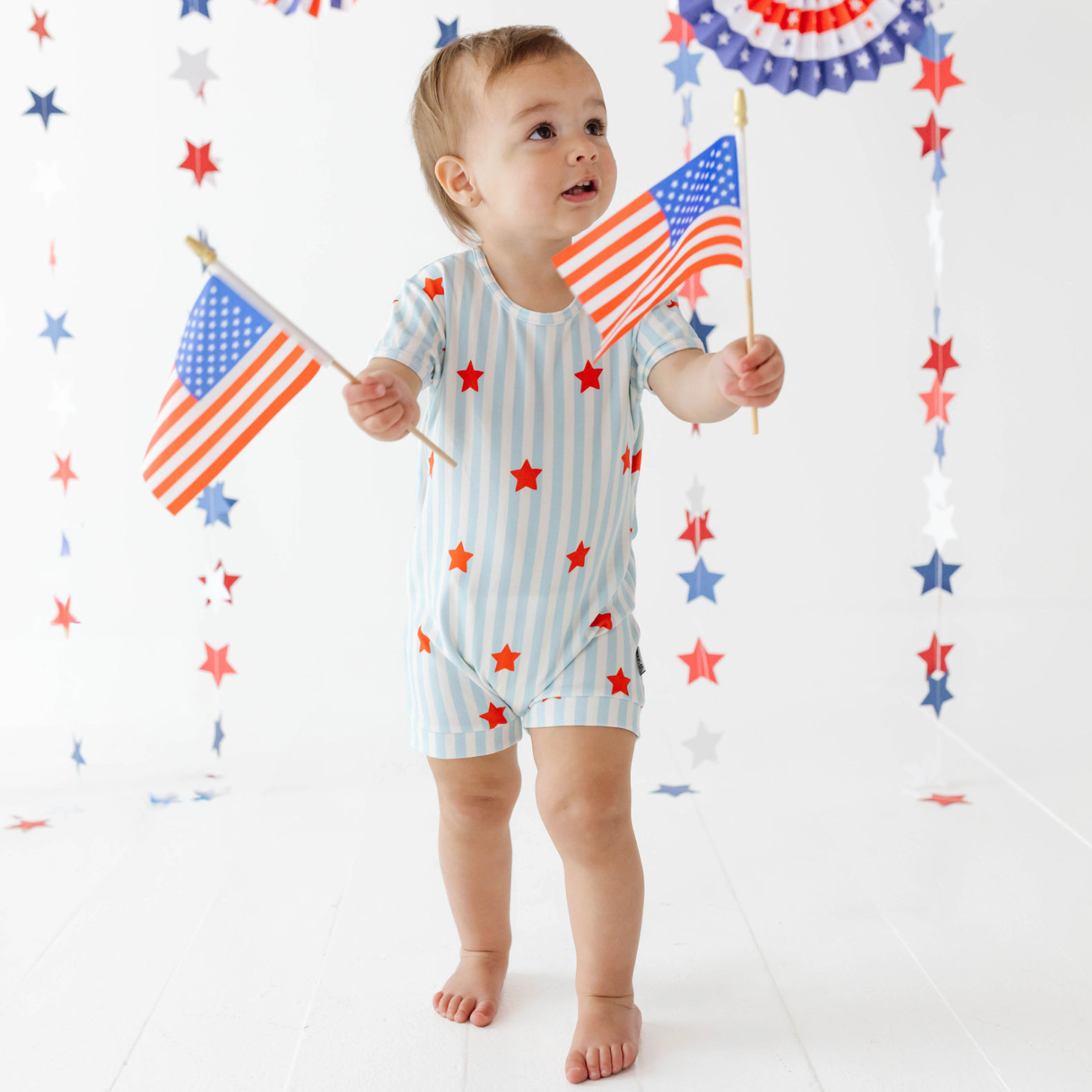 Child wearing a onesie with star pattern, holding American flags against a decorative background.
