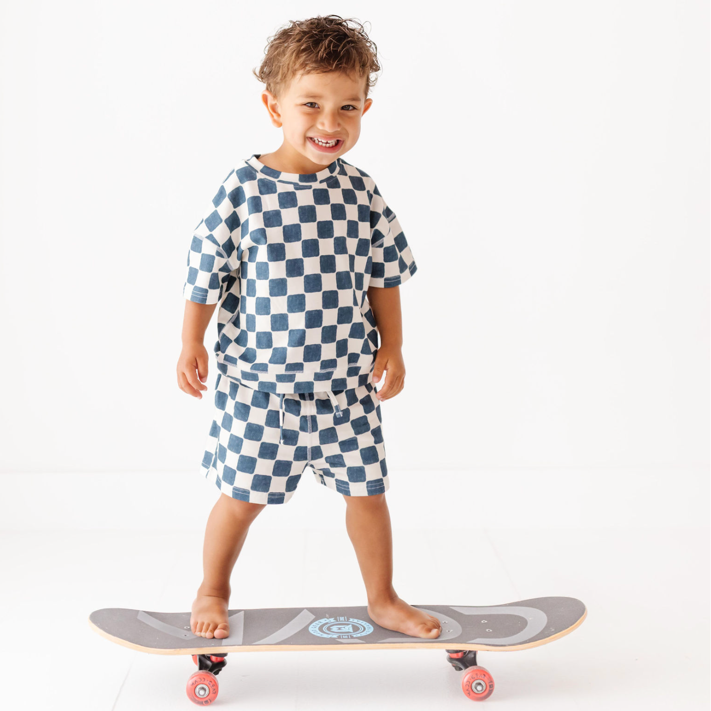 Child wearing a blue and white checkered outfit standing on a skateboard.