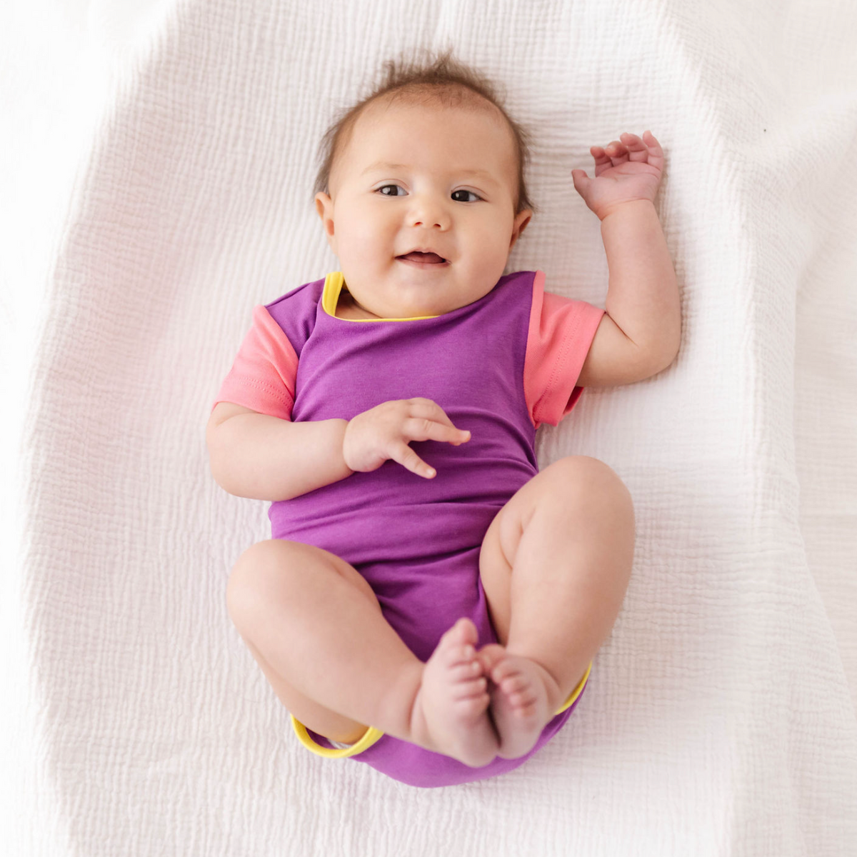 Baby wearing a purple and pink onesie lying on a white blanket
