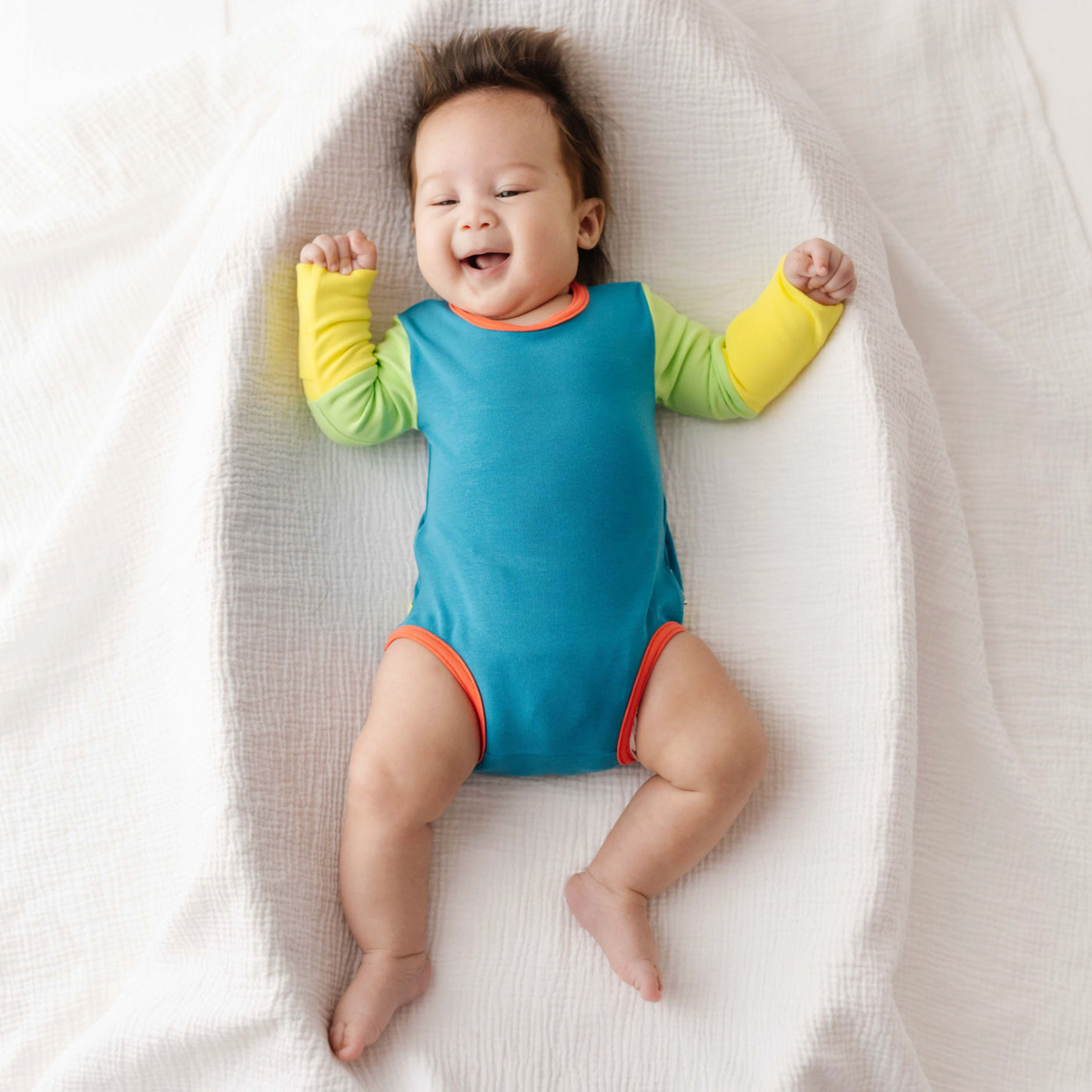 Baby lying on a white blanket wearing a colorful onesie.