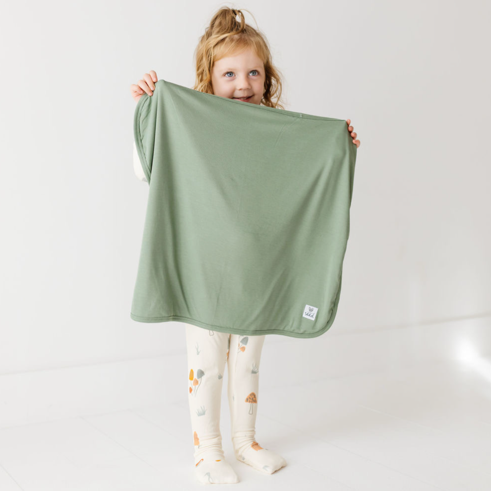 A young child with blonde hair in a ponytail holds up a sage green blanket or throw against a plain white studio background. The child is wearing white tights with a small animal print.