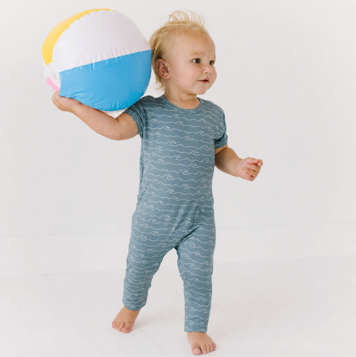 A toddler standing barefoot and holding a beach ball, wearing a short-sleeve one-piece jumpsuit in muted blue-gray with a white wavy line pattern. The background is a clean white studio setting.
