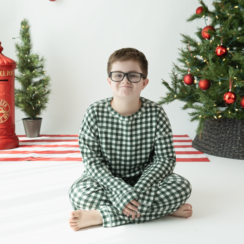 A young boy wearing green and white gingham pajamas sits cross-legged on a white floor in a decorated Christmas room, with a Christmas tree, red ornaments, a small red fire hydrant decoration, and a potted tree in the background.