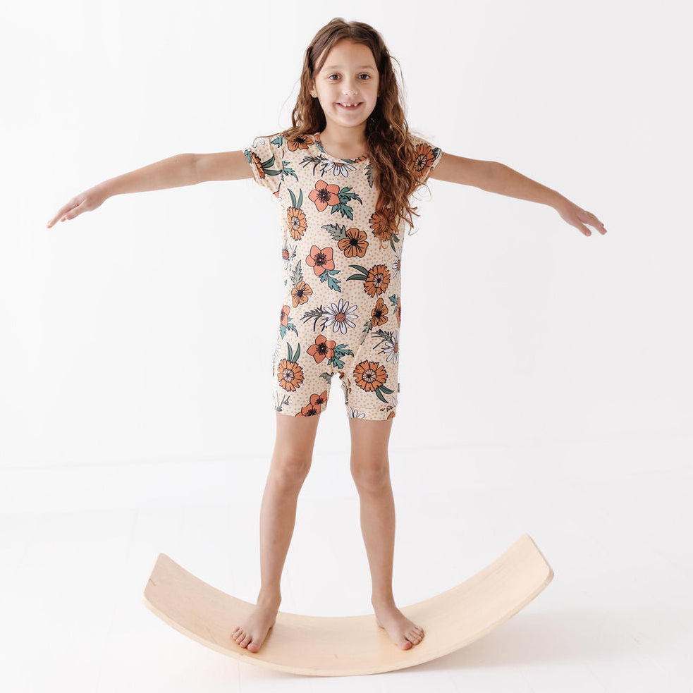 Young girl in a floral outfit standing on a wooden balance board against a white background