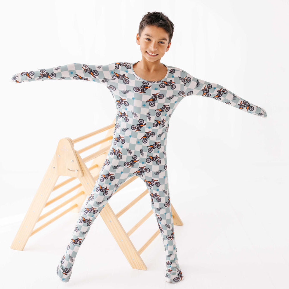 Child wearing a patterned onesie standing next to a wooden stool on a white background