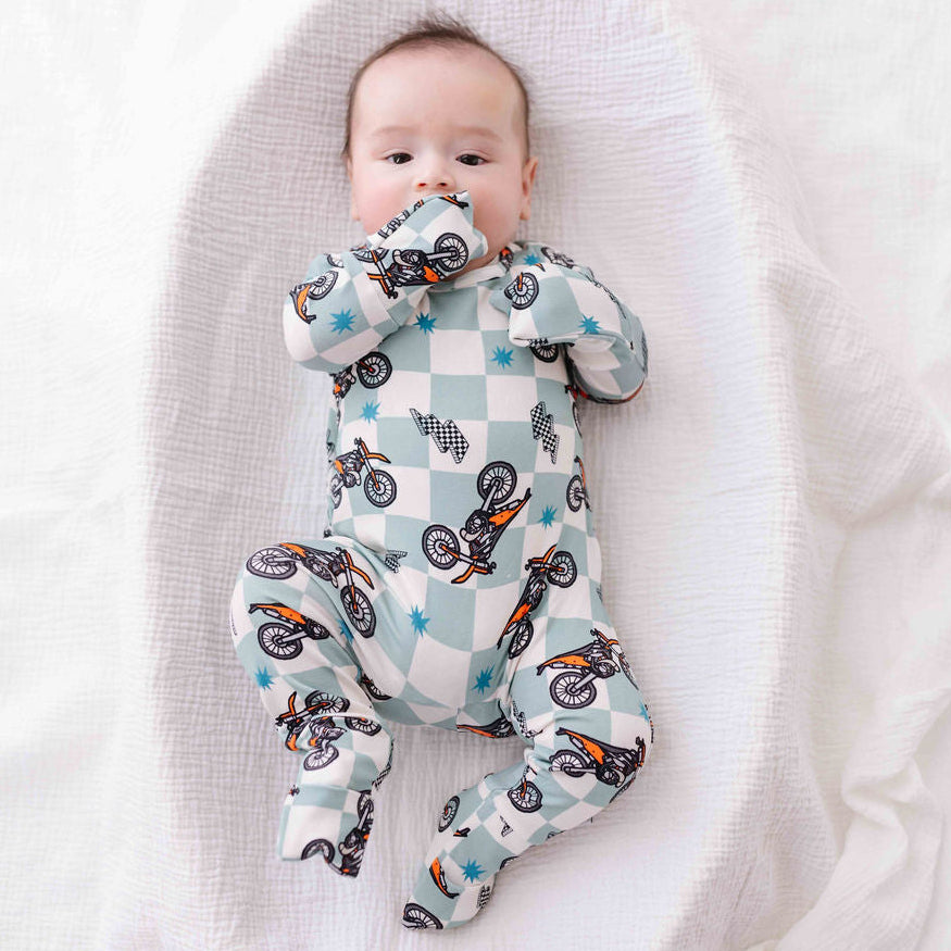 Baby lying on a white blanket wearing a checkered onesie with bike designs.