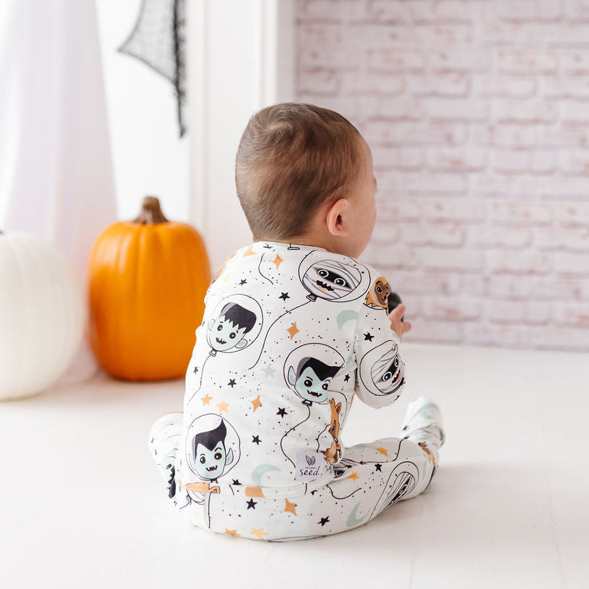 A baby sits on a white floor facing a decorated fireplace, wearing a white footed sleeper with a cartoon astronaut print. A white pumpkin, an orange pumpkin, and a ghost decoration are visible in the background.