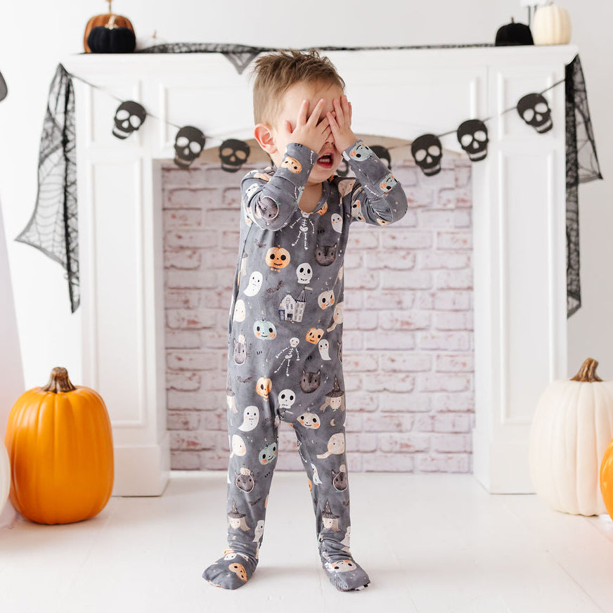 A toddler stands in front of a white fireplace wearing a gray full-body Halloween pajama with a ghost and skull print. The room is decorated with pumpkins, black lace, and ghost props.