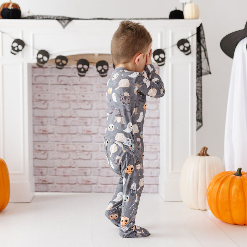 A toddler stands in front of a white fireplace wearing a gray long-sleeve pajama top and matching footed pajama bottoms with a Halloween print of ghosts, skulls, and pumpkins. The room is decorated for Halloween with a ghost figure, pumpkins, black spiderweb bunting, and skull garland.