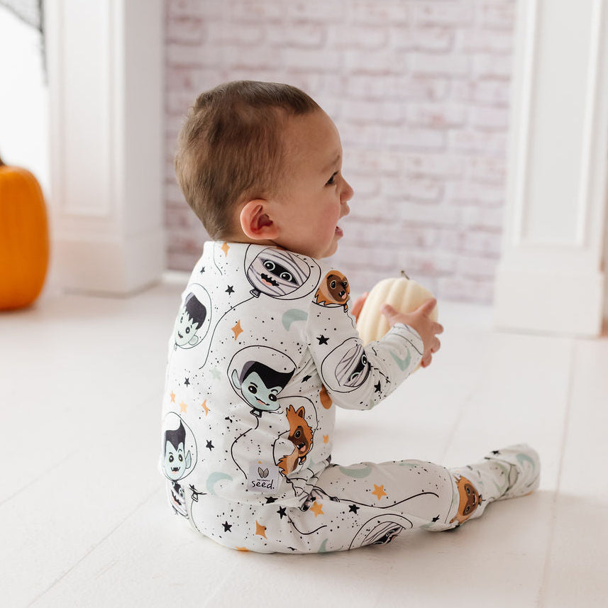 A baby sitting on a white floor in a decorated room, wearing a white footed sleeper with a cartoon space-themed print featuring black cats, orange stars, and gray planets.