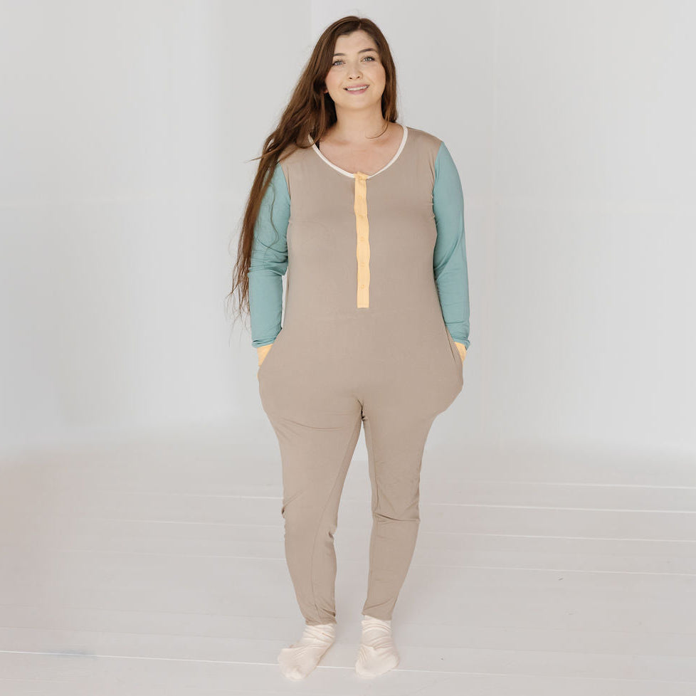 A woman modeling a beige color-blocked jumpsuit with sage green long sleeves and a pale yellow front placket, set against a bright white studio background.