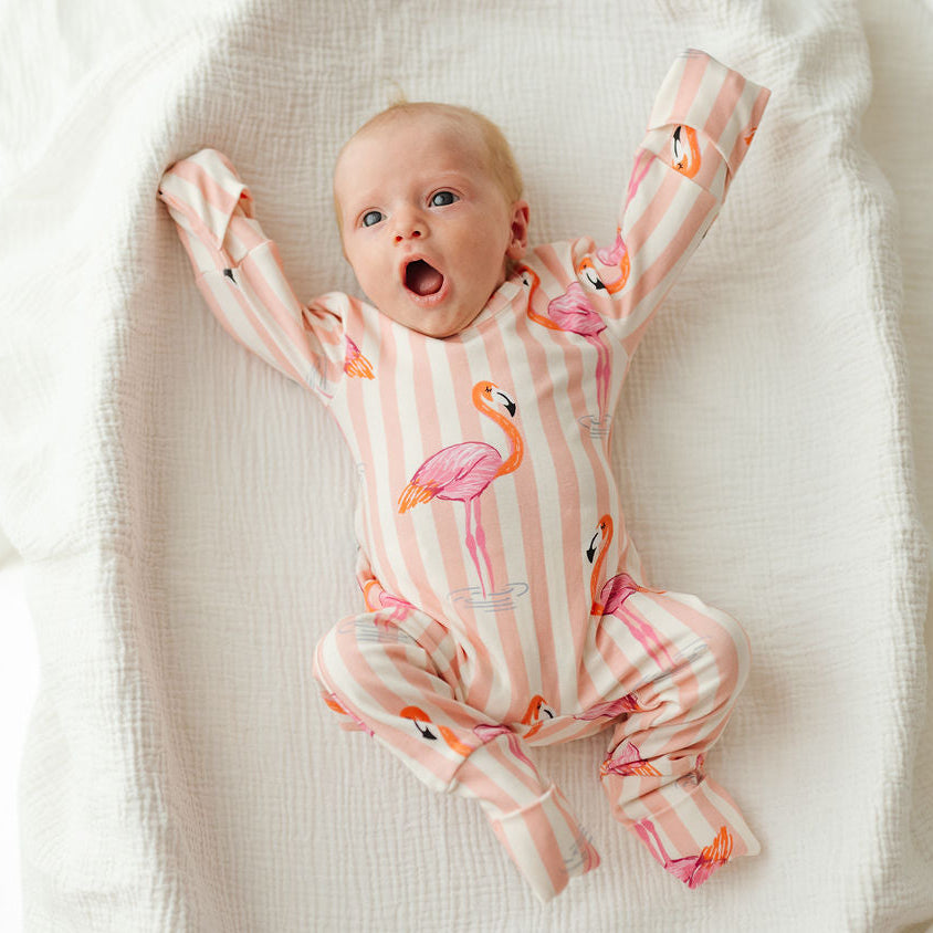 A baby lying on a textured white muslin blanket, wearing a pink and white striped footed sleeper with a flamingo print.