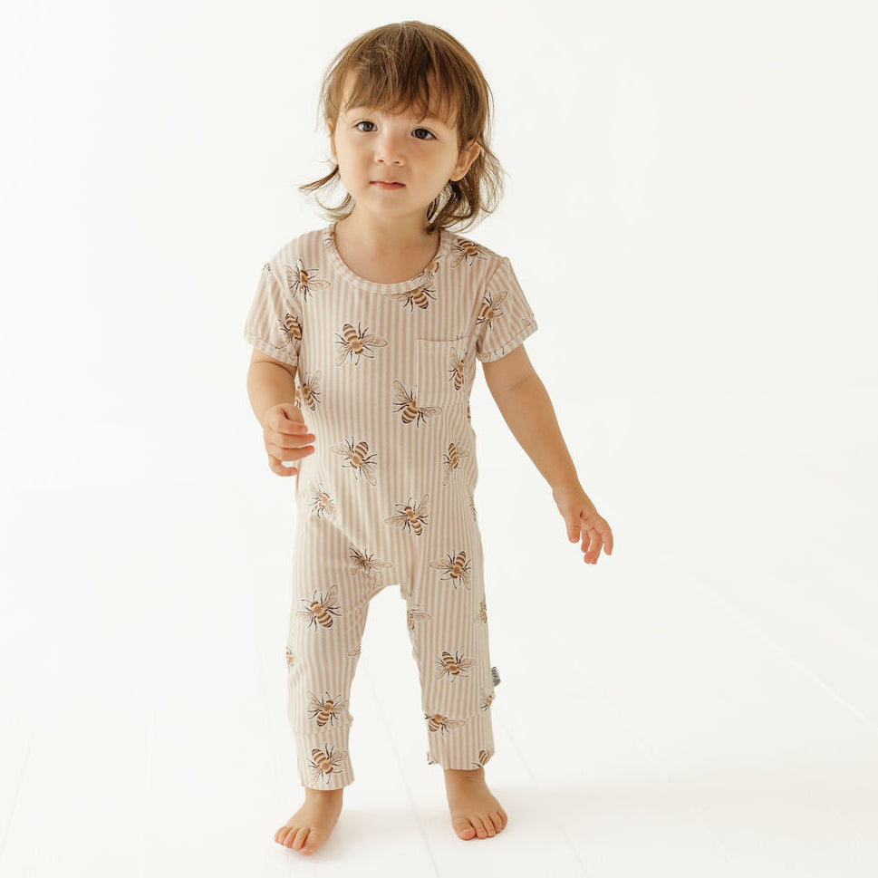 A toddler standing barefoot against a plain white studio background, wearing a short-sleeve one-piece jumpsuit with vertical beige and white stripes and a scattered bee print.
