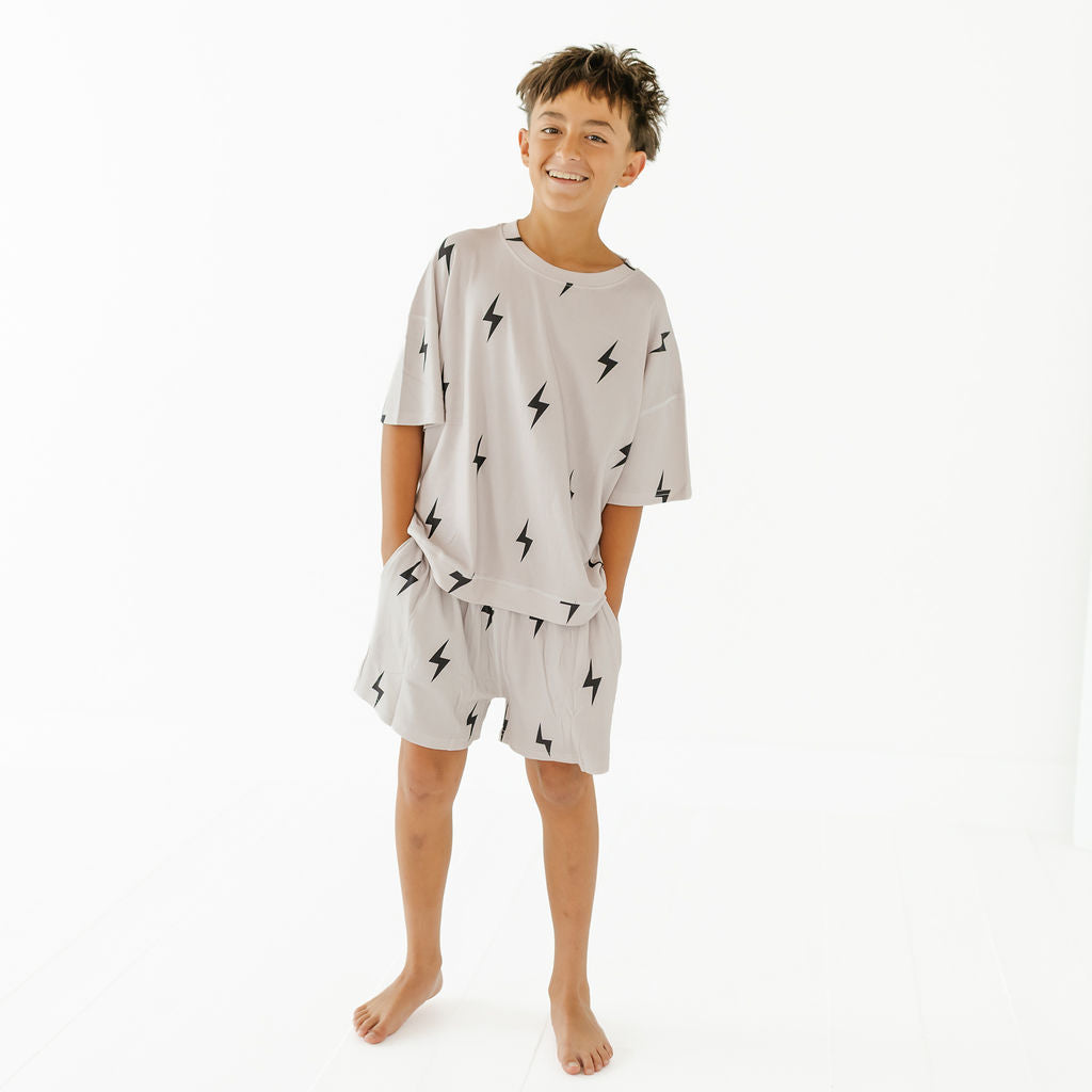 A young boy stands barefoot against a plain white studio background, wearing a light beige or off-white short-sleeve pajama set with a black lightning bolt print. The set includes a loose T-shirt and matching shorts.
