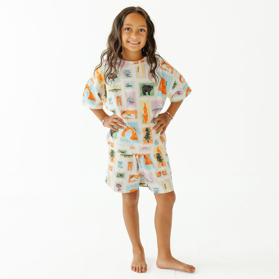 A young girl stands barefoot against a plain white studio background, wearing a short-sleeve matching set with a light blue base and a colorful illustrated print of campfire scenes, trees, and animals.
