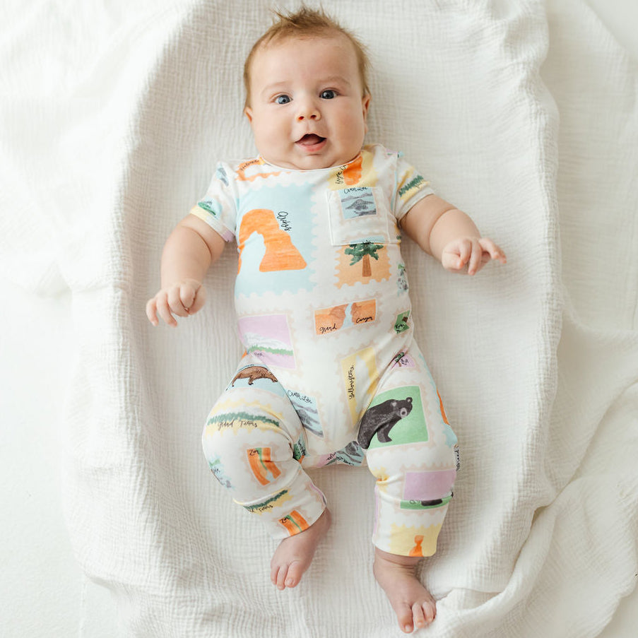 A baby lying on a white textured blanket, wearing a short-sleeve one-piece jumpsuit with a colorful patchwork print of animals and landscape motifs.