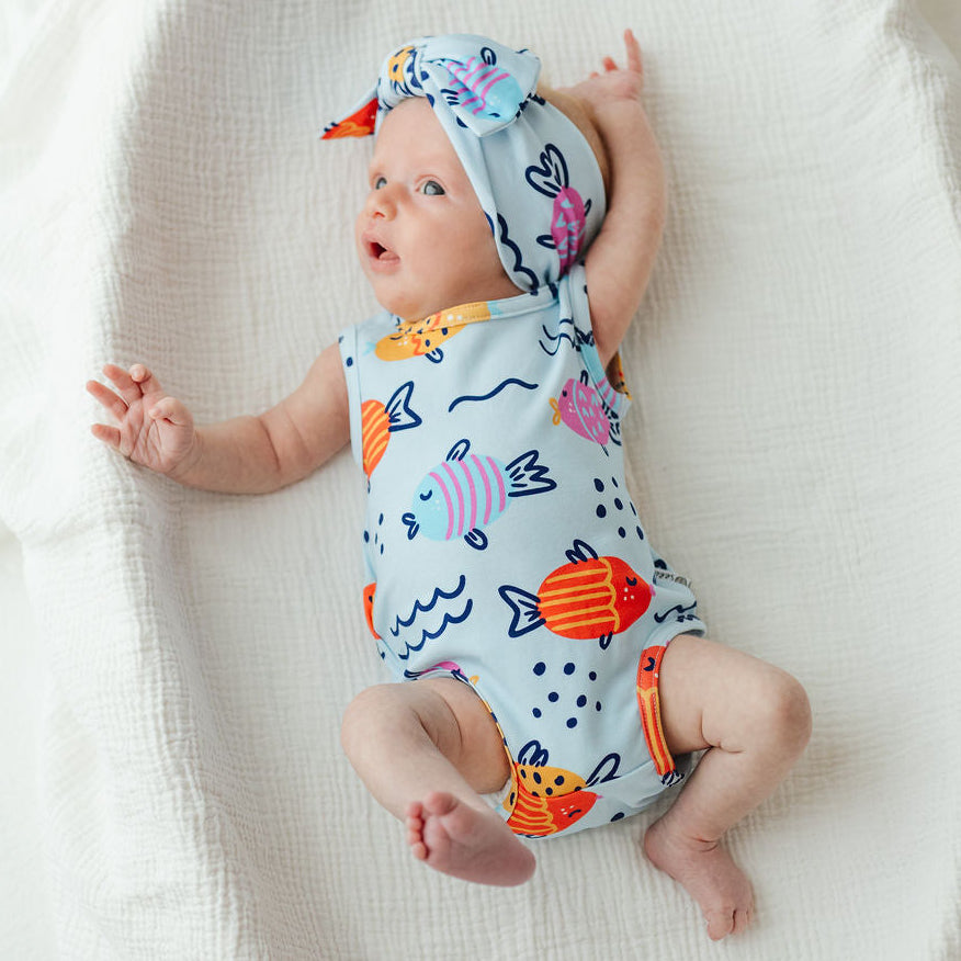 A baby lying on a white textured blanket, wearing a light blue one-piece swimsuit and matching hat with colorful fish and wave patterns.
