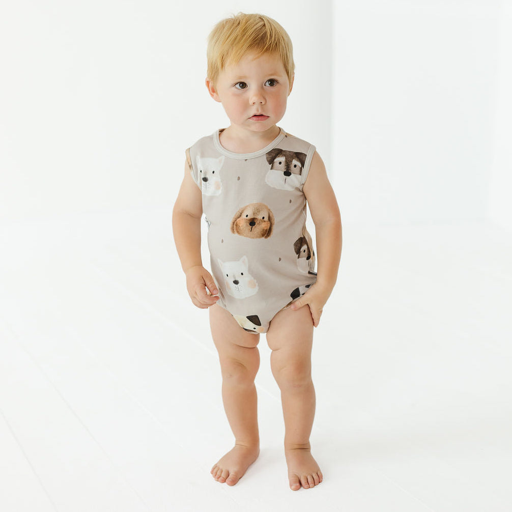 A toddler standing barefoot against a clean white studio background, wearing a sleeveless beige bodysuit printed with dog faces.