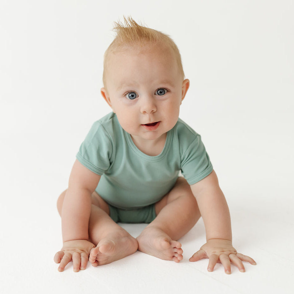 A baby with light hair and blue eyes sits on a white studio floor wearing a sage green short-sleeve bodysuit.