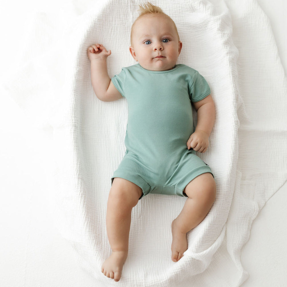 A baby lying on a white textured blanket, wearing a sage green short-sleeve romper.