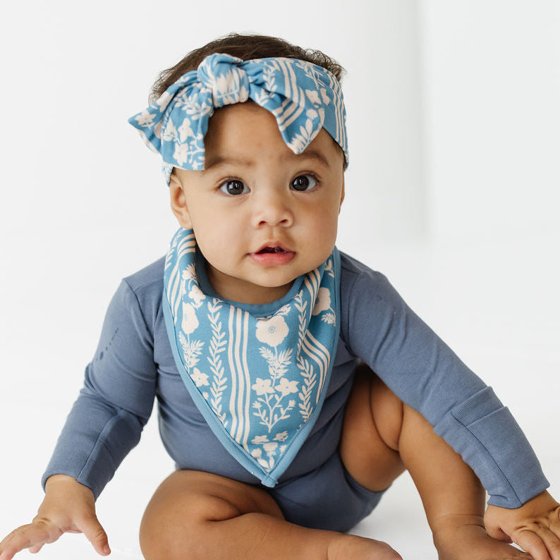 A baby wearing a slate blue long-sleeve bodysuit, a matching blue and white patterned bib, and a matching blue and white patterned headband, sitting on a white studio floor.