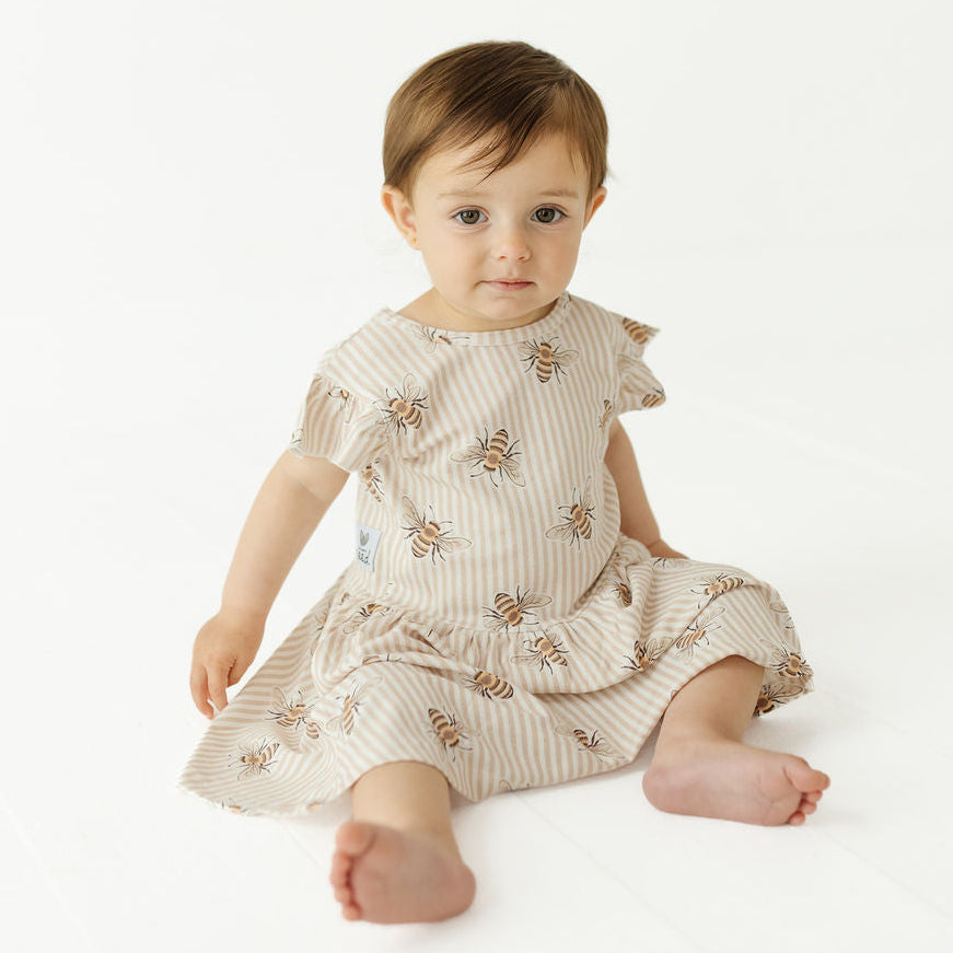 A baby sitting on a white studio floor wearing a short-sleeve cream dress with thin vertical stripes and embroidered bees.