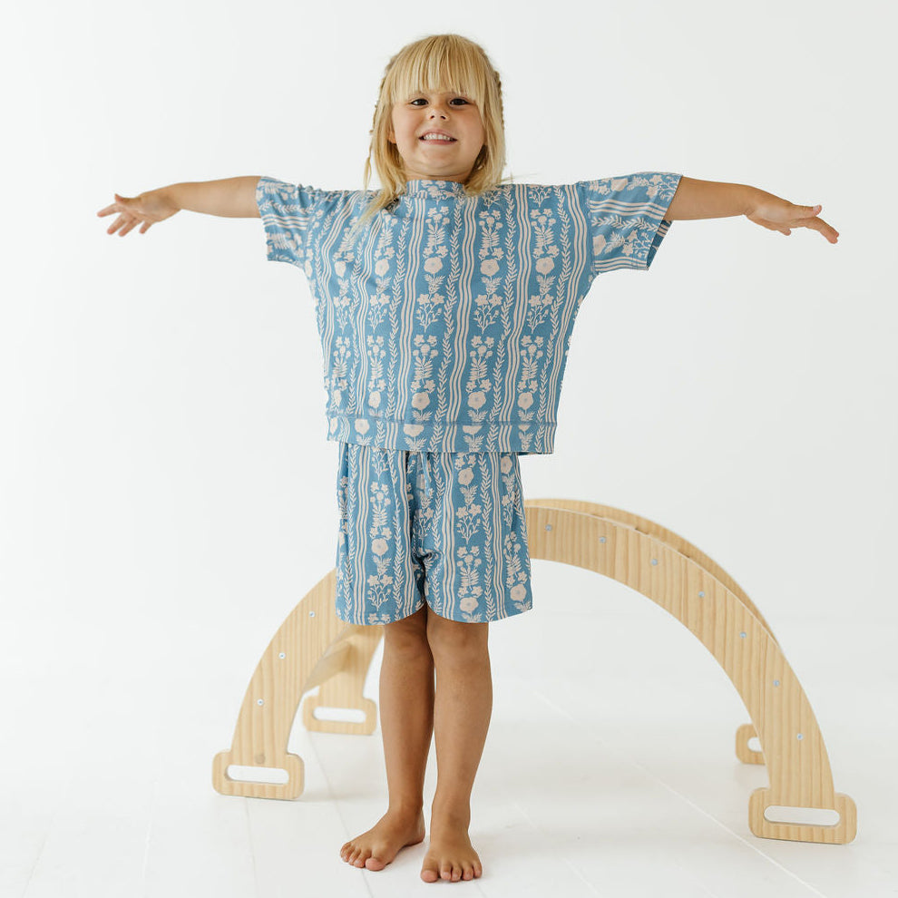 A young child stands barefoot with arms outstretched, wearing a blue and white patterned short-sleeve top and matching shorts, posed in front of a light wood arch on a white studio background.