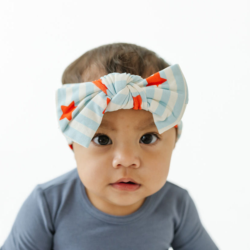 A baby wearing a light blue and white striped bow headband with orange star accents, paired with a slate blue long-sleeve shirt, photographed against a solid white background.